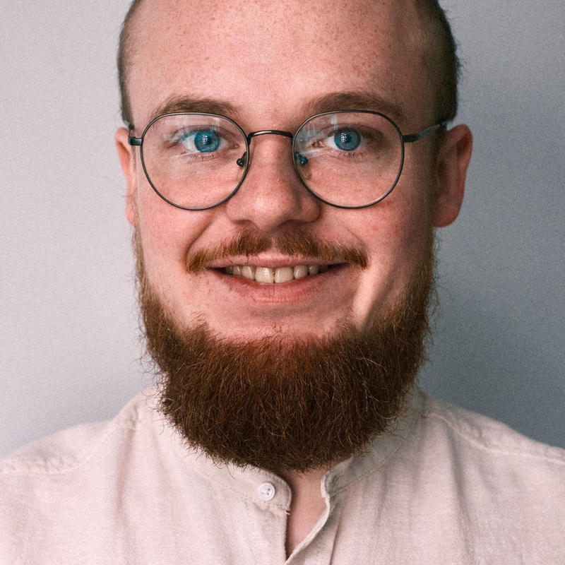 Frontal portrait: smiling person with short hair, round metal glasses, and a reddish full beard; wearing a linen shirt against a light background.